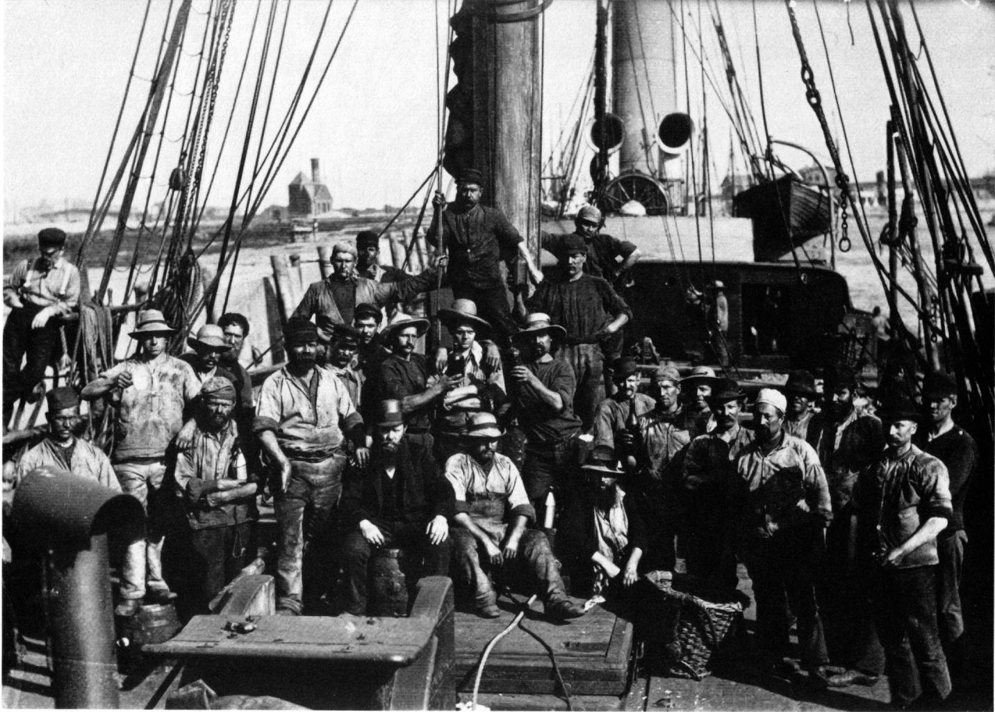 Schauerleute mit ihrem »Vizen« (auf der Tonne sitzend). Das Foto zeigt sogenannte »schwarze Schauerleute« – die Männer arbeiteten als Kohlenträger (Hamburger Hafenstreik, 1899).