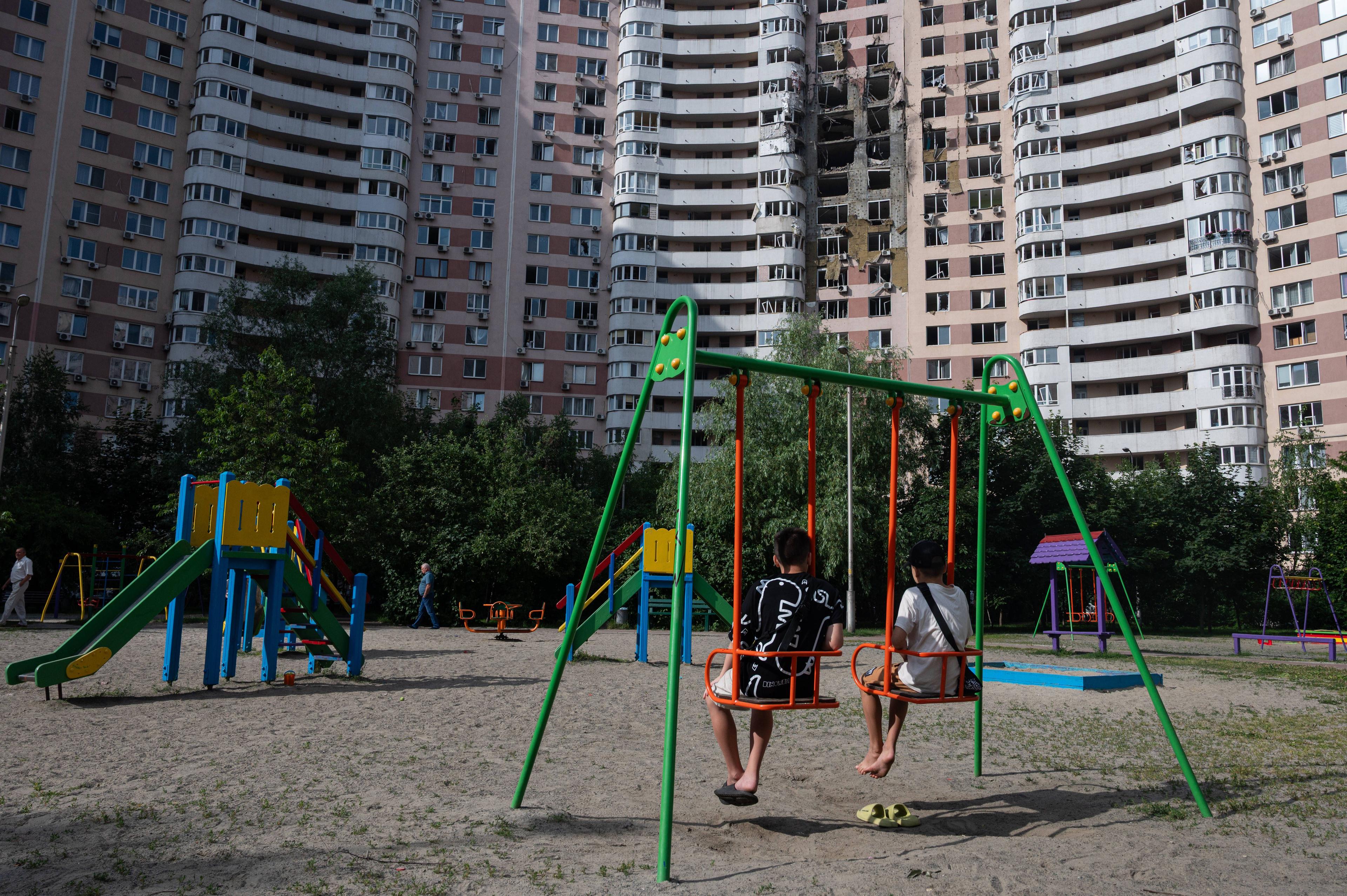 Zwei Kinder auf einem Spielplatz in Kiev, dahinter ein durch eine russische Rakete beschädigtes Wohngebäude.
